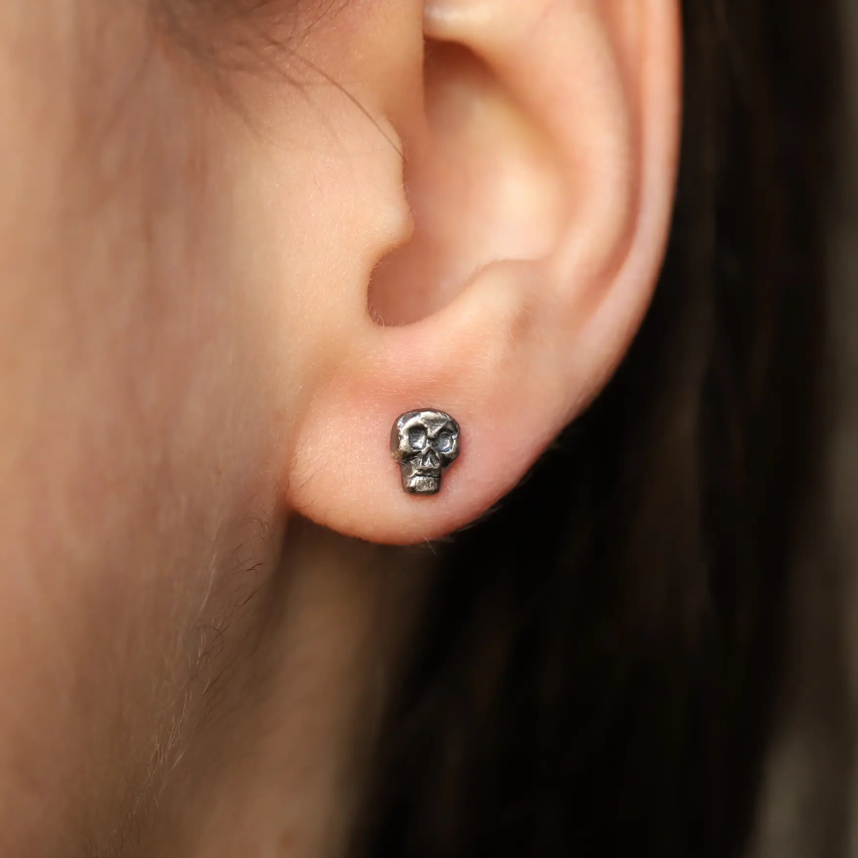 Oxidized Silver Skull Stud Earrings worn on ear, handmade in sterling silver 925, dark oxidized finish, unisex jewelry, 5mm x 6mm.