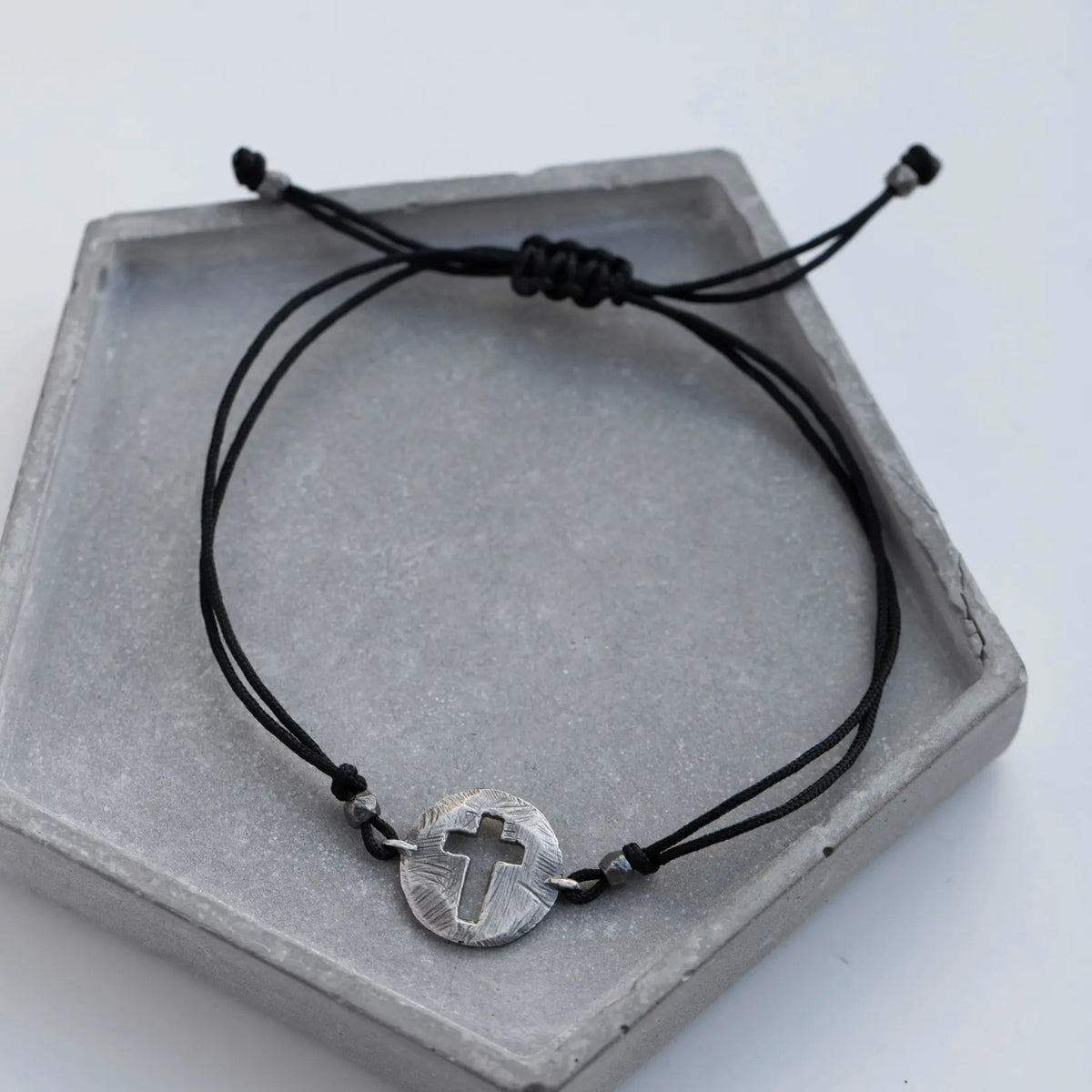 Adjustable oval cross bracelet with black cord on gray display tray