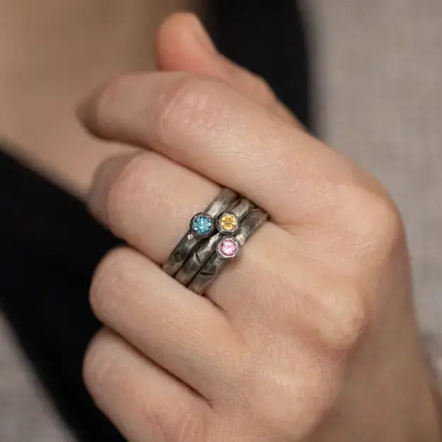 Hand wearing stack of oxidized silver rings with multicolored gemstones, showcasing unique brutalist design and artistry.